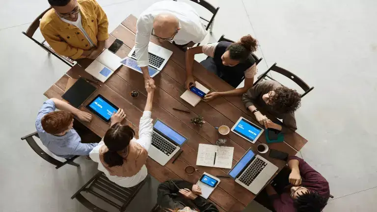 Engaged core community article link & header image - Top-down view of group of colleuges sitting around a wooden table interaacting with one another while working on laptops and mobile devices.